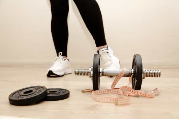 Lower body shot of a person in workout attire standing near a disassembled dumbbell and a measuring tape on a wooden floor, highlighting tools used for strength training and fitness monitoring.